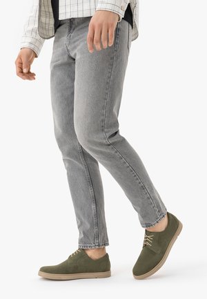 Muž v šedých džínách, zelených šněrovacích botách a bílé kostkované košili kráčí před jednobarevným bílým pozadím.