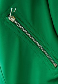 Tessuto in ecopelle verde con una zip metallica visibile, caratterizzata da denti e cursore argentati, bordato da cuciture decorative.