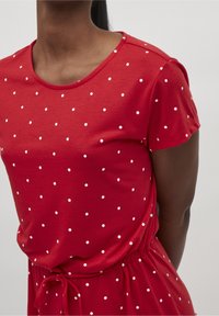 Punainen t-paita pyöreällä kaula-aukolla, lyhyillä hihoilla ja valkoisilla pilkuilla. Yhdessä on kireä vyötärö, jossa on nauhatieto. Sileä pinta.