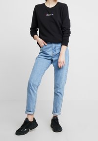 Felpa nera con logo "WRANGLER", jeans in denim blu chiaro e sneakers nere. Abbigliamento casual complessivo con orli arrotolati sui jeans.