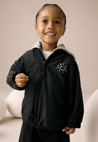 Veste noire à fermeture éclair avec une capuche grise, arborant un motif de soleil souriant en blanc sur le côté gauche. Tissu lisse avec des poignets froncés.