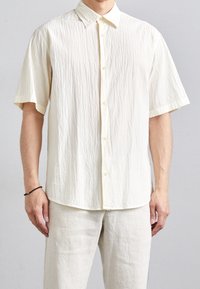 Lyhythihainen, nappikiinnitteinen paita vaaleanvalkoisena, jossa on teksturoitu kangas hienovaraisella rypistymisellä ja klassisella kauluskotelolla.