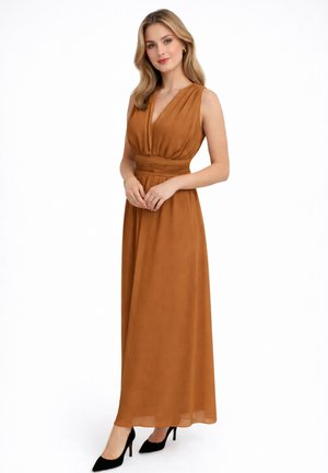 Donna con capelli chiari che indossa un abito lungo senza maniche color ruggine e tacchi alti neri, in piedi con le mani leggermente intrecciate.