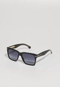 Óculos de sol quadrados pretos com lentes escuras em degradê, armação em plástico, detalhes em dourado nas laterais. Logotipo da marca na haste.