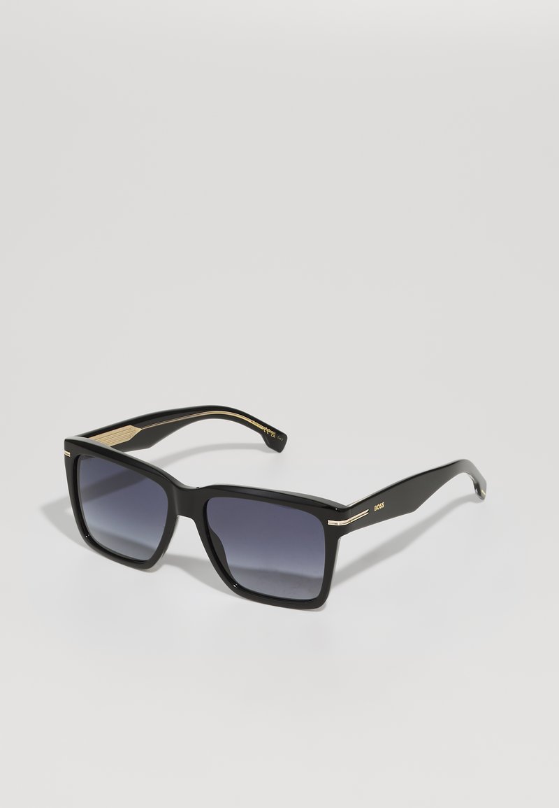 Óculos de sol quadrados pretos com lentes escuras em degradê, armação em plástico, detalhes em dourado nas laterais. Logotipo da marca na haste.