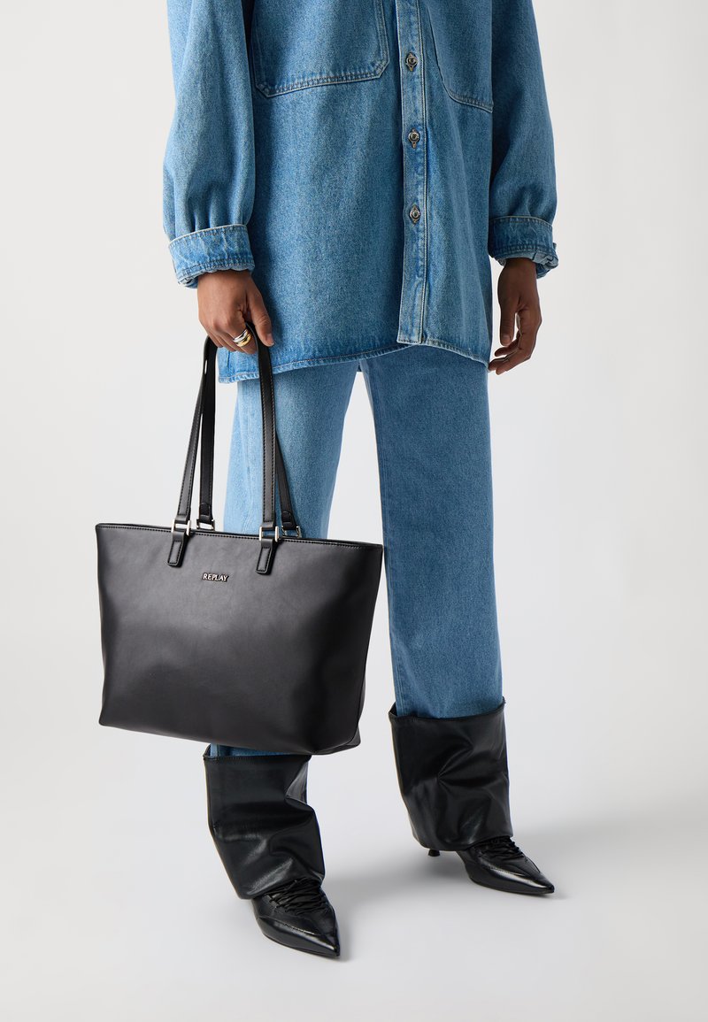 Svart lær totebag med to skulderstropper, rektangulær form, preget logo og glatt tekstur. Brukt med et denimantrekk og spisse støvler.