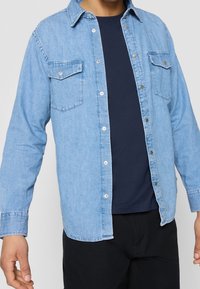 Camicia in denim azzurro chiaro con maniche lunghe, frontale con bottoni, due tasche sul petto e colletto; indossata sopra una t-shirt girocollo blu navy.
