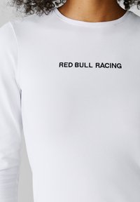Kobieta ubrana w białą koszulę z długim rękawem z czarnym haftowanym napisem „RED BULL RACING” na piersi.