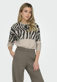 Jersey de punto con un cuerpo beige y rayas de cebra negras, cuello redondo acanalado y mangas largas. Combinado con pantalones de talle alto en color verde oliva.