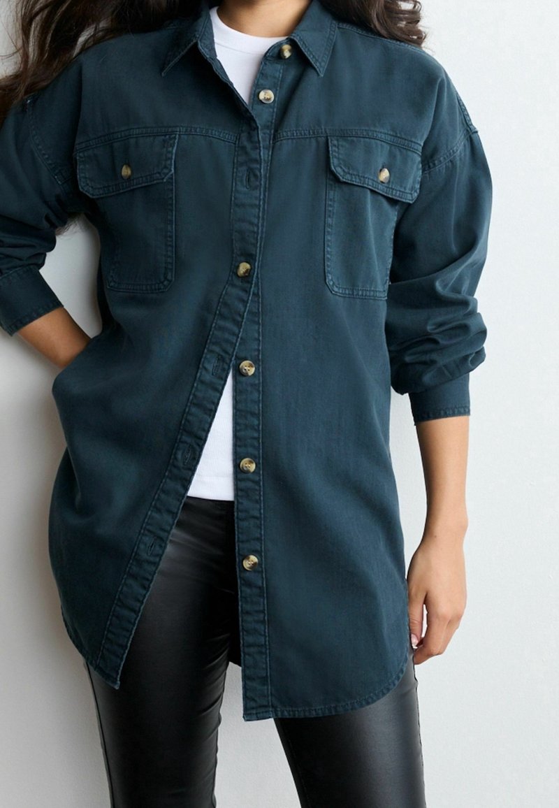 Femme portant une chemise bleu foncé à manches longues boutonnée sur un haut blanc et un pantalon en cuir noir, avec les manches retroussées sur un bras.