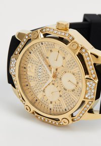 Montre-bracelet Guess en or avec un cadran studé de cristaux, trois sous-cadrans pour le jour, la date et l'heure au format 24 heures, et un bracelet noir.