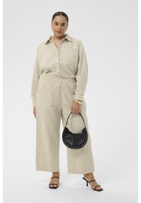 Femme portant une chemise beige à rayures et un pantalon à jambes larges, tenant un sac à main noir, debout avec des sandales noires à talons.