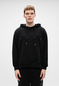 Sudadera con capucha negra que presenta un bolsillo frontal, cuerdas ajustables y la palabra "HUGO" impresa en blanco en el pecho. Textura suave y uniforme.
