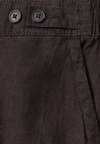 Gros plan d’un pantalon en tissu foncé montrant la ceinture avec deux boutons noirs et des détails de couture visibles.