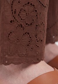 Brown fabric with floral cut-out embroidery on pant leg above a foot wearing a tan leather shoe.