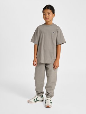 Ung person iført en løs grå T-shirt, matchende træningsbukser og hvide sneakers med grønne accenter på hvid baggrund.