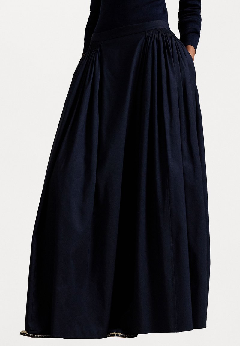 Sieviete, kas valkā garu, pilnu, tumši zilu svārku ar savilktām krokām jostas vietā un rokām ieliktām sānu kabatās.