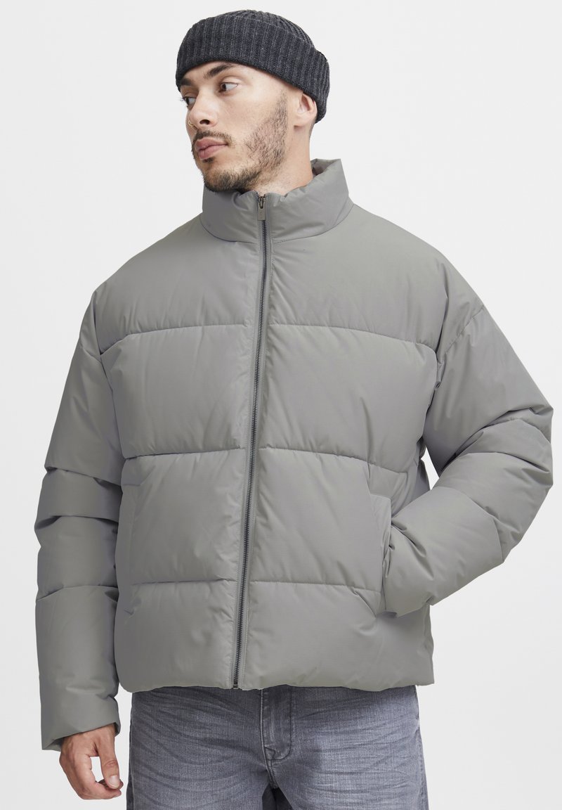 Veste matelassée grise claire avec col montant, fermeture éclair intégrale et poches latérales. Texture lisse avec un motif matelassé horizontal.