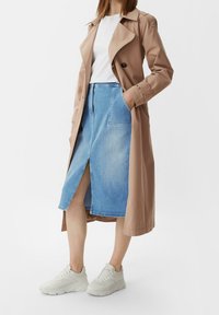 Kobieta ubrana w beżowy trencz, biały top, jasnoniebieską spódnicę dżinsową z przodu rozcięciem i białe chunky sneakers, stojąca na tle białym.