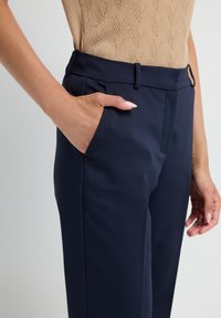 Kvinna i marinblå byxor med handen i fickan och en beige texturerad ärmlös topp mot en enfärgad bakgrund.