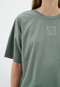 Olivově zelené oversized tričko vyrobené z hladkého materiálu, se čtvercovým logem s tečkovaným obrysem na oblasti hrudníku a krátkými rukávy.