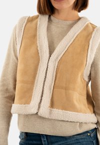 Gilet en daim beige avec une douce bordure en shearling crème le long des bords, présentant un décolleté en V profond et un design sans manches. Porté par-dessus un pull léger.