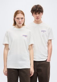 Νεαρός άνδρας και γυναίκα φορούν λευκά t-shirts με το λογότυπο Tommy και σκούρα καφέ παντελόνια, στέκονται πλάι-πλάι μπροστά σε απλό φόντο.