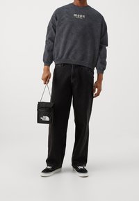 Sudadera gris oscuro con logotipo de "luna", pantalones vaqueros negros holgados y zapatillas negras. Lleva una pequeña bolsa negra con acentos de logotipo en blanco.