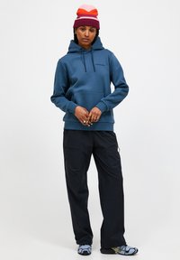 Blauer Hoodie mit Kordelzug, vorderer Tasche und Logo; kombiniert mit schwarzen Cargo-Hosen; akzentuiert durch eine farbenfrohe gestreifte Strickmütze.