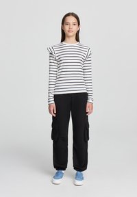 Pantaloni cargo neri con tasche laterali abbinati a una camicia a maniche lunghe bianca con righe e dettagli arricciati sulle spalle. Sneakers blu completano il look.