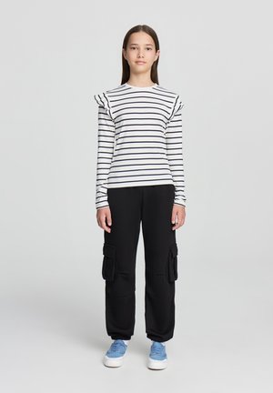 Pantaloni cargo neri con tasche laterali abbinati a una camicia a maniche lunghe bianca con righe e dettagli arricciati sulle spalle. Sneakers blu completano il look.