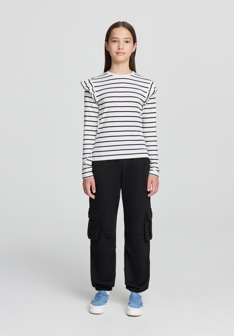 Pantaloni cargo neri con tasche laterali abbinati a una camicia a maniche lunghe bianca con righe e dettagli arricciati sulle spalle. Sneakers blu completano il look.