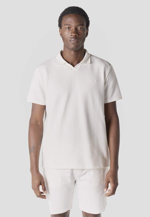 Muž stojaci čelom vpred, oblečený v bielej textúrovanej polokošeli s krátkym rukávom a zodpovedajúcich kraťasoch na jednoduchom svetlom pozadí.