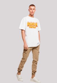Weißes T-Shirt mit orangefarbenem "Stranger Things"-Logo, khakifarbene Cargo-Hosen mit Seitentaschen und weiße Sneaker. Lässiger Schnitt und Design.