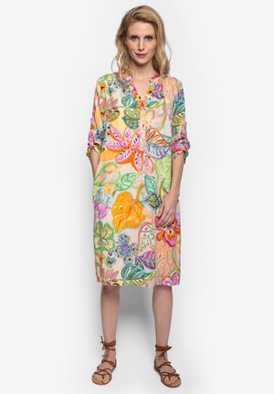 Vrouw die een kleurrijke jurk tot op de knie draagt met opgerolde mouwen en patronen van bladeren en bloemen, staand op bruine sandalen met bandjes.