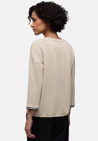 Frau mit lockigem Haar, die ein beiges Langarmshirt mit schwarz gestreiften Bündchen und schwarze Hosen trägt, steht im Profil vor einem schlichten Hintergrund.