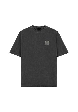 Man staat met het gezicht naar voren, draagt een vervaagde zwarte oversized t-shirt, lichtgroene spijkerbroek en zwart-witte sneakers tegen een effen grijze achtergrond.