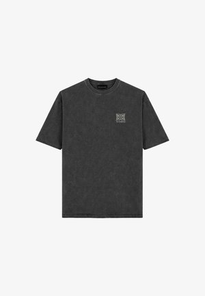Ein Mann steht frontal, trägt ein ausgewaschenes schwarzes oversized T-Shirt, hellgrüne Jeans und schwarz-weiße Turnschuhe vor einem einfachen grauen Hintergrund.