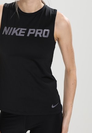 Frau trägt ein ärmelloses schwarzes Nike Pro Top mit grauem Logo-Text und kleinem Swoosh nahe dem Saum, steht vor einem einfarbigen Hintergrund.