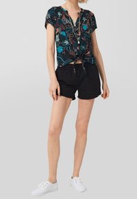 Top floreale a maniche corte in turchese e nero, annodato davanti, abbinato a pantaloni corti neri e scarpe da ginnastica bianche. Design semplice e casual.