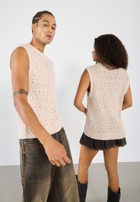 Pull sans manches en tricot beige clair, présentant un tissage ouvert texturé et un ourlet côtelé, associé à un jean foncé et une jupe évasée.