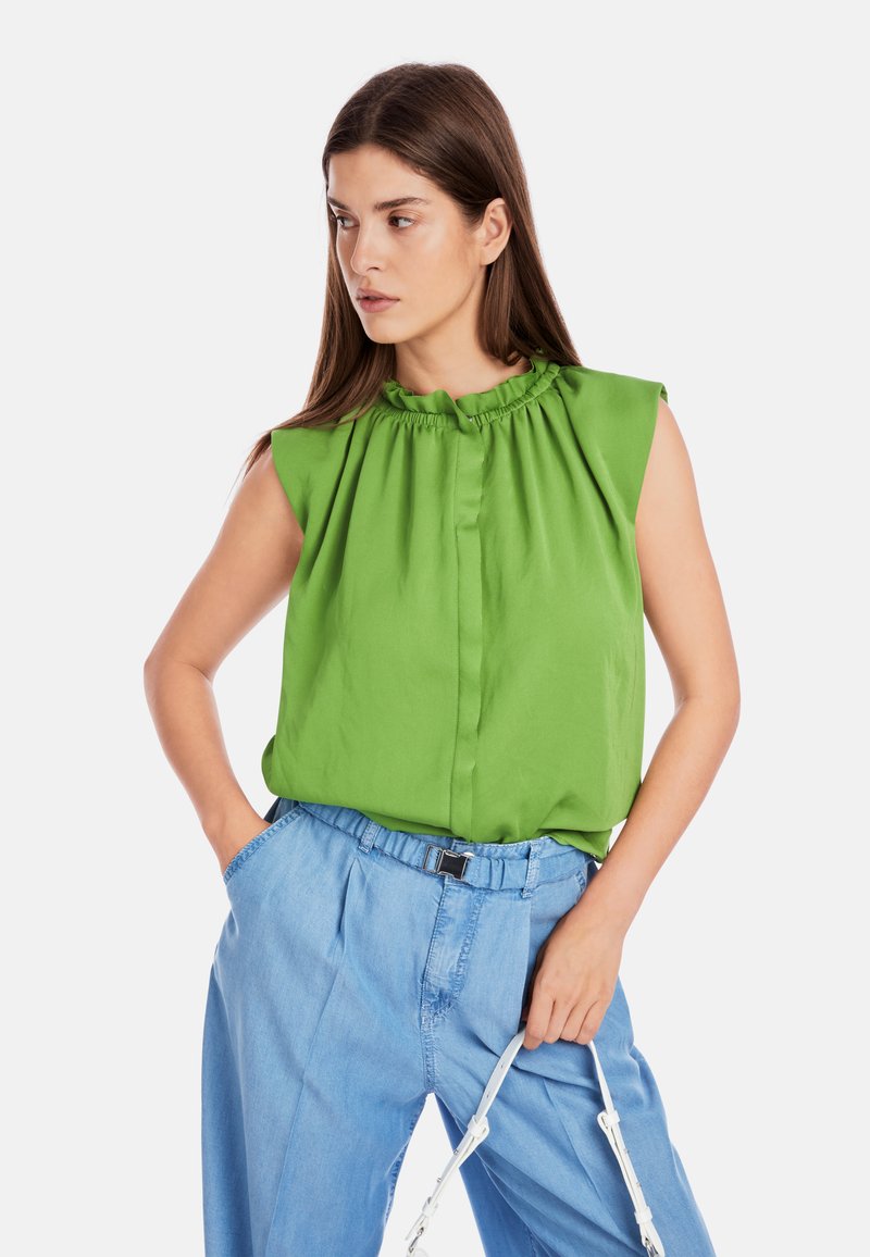 Grüne ärmellose Bluse mit gerafftem Ausschnitt und lässigem Schnitt, kombiniert mit hellblauen hochgeschnittenen Hosen mit vorderenFalten.