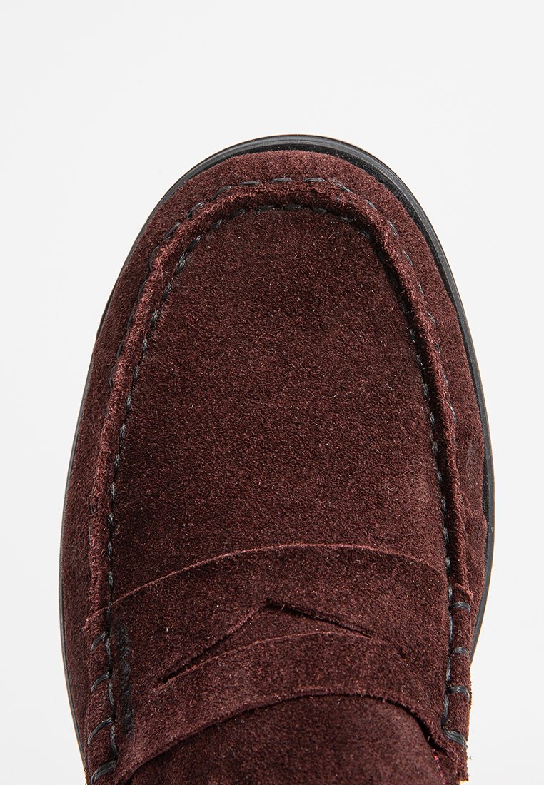 Mocassino in suede marrone con cuciture visibili e dettaglio a striscia penny sopra, su uno sfondo bianco.