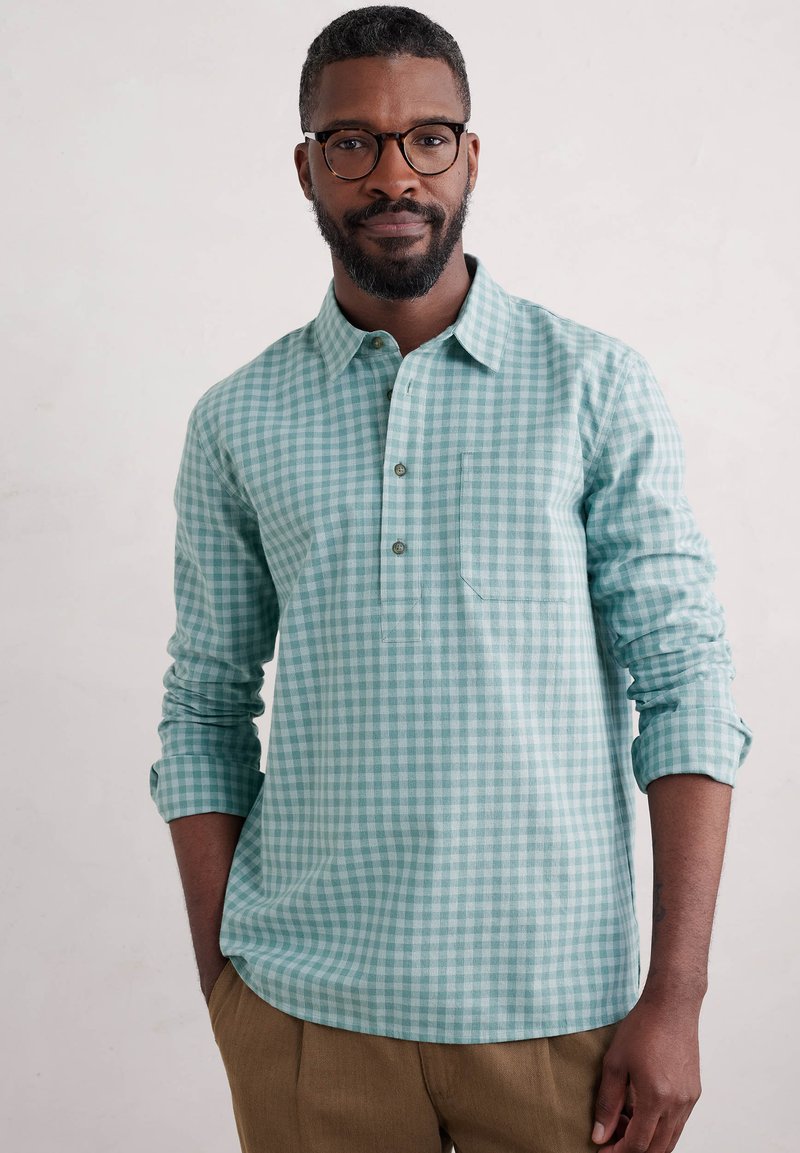 Chemise à carreaux bleu clair et blanche, à manches longues, avec un col et un bouton sur le devant, dotée d'une poche poitrine. Portée avec un pantalon khaki.