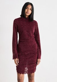 Burgundin pitsire dressi pitkien hihojen kanssa ja tiukassa siluetissa. Teksturoitu kuvio ja laineileva helma, jossa on rypyttyjä yksityiskohtia.