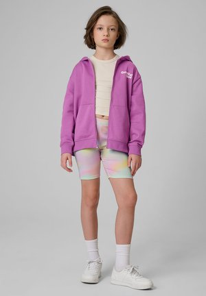Dziecko stoi, ubrane w fioletową bluzę z kapturem i suwakiem, krótki beżowy top, pastelowe spodenki rowerowe tie-dye, białe skarpetki i białe trampki na szarym tle.