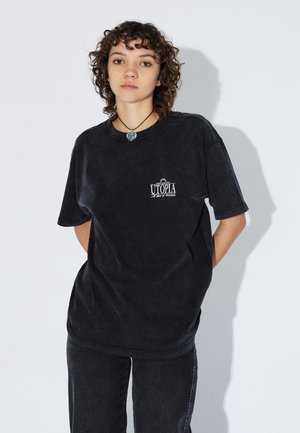 Zwart katoenen T-shirt met een losse pasvorm, korte mouwen en een grafiek op de borst met de tekst "UTOPIA." Gladde textuur met een vervaagde uitstraling.