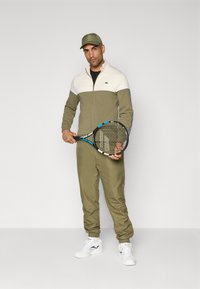 Olijfgroene trainingsoutfit met crèmewitte bovenpaneel, ritsluiting, elastische manchetten en bijpassende pet; houdt een tennisracket vast. Witte sneakers.