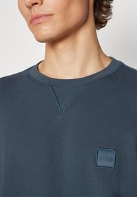 Jeune personne portant un sweat-shirt bleu marine à col rond avec un petit patch carré du logo BOSS sur la poitrine, regardant vers la gauche.
