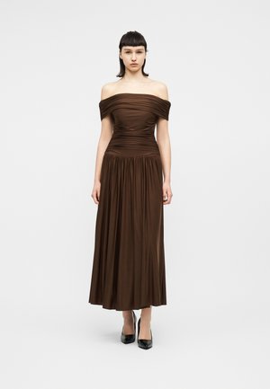 Frau steht aufrecht und trägt ein braunes schulterfreies Kleid mit Raffungen und einem plissierten Rock sowie schwarze spitze Absatzschuhe vor einem schlichten Hintergrund.